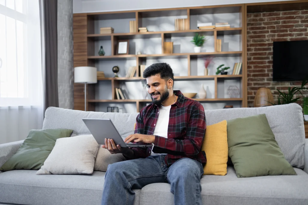 smiling young man sitting on a gray sofa using a laptop in a modern living room with a bookshelf and brick wall background.
