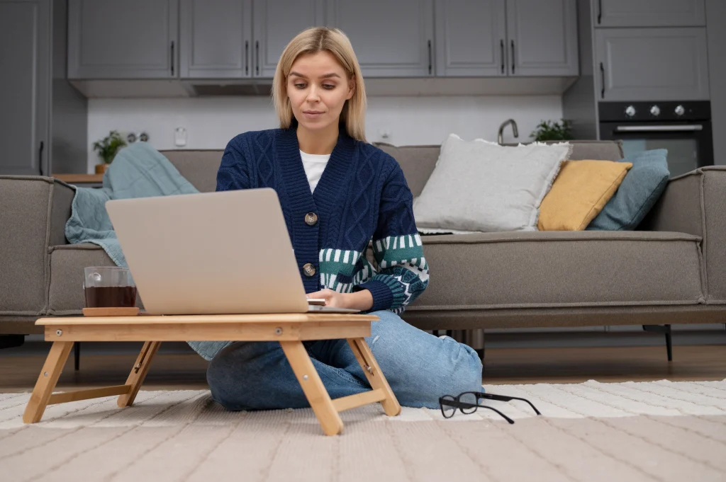A woman working on a laptop placed on a wooden bed desk while sitting on a rug in a minimalist home office or kitchen setting.