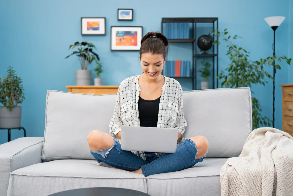 A happy young woman sitting cross-legged on a sofa while typing on a laptop, with a blue wall and indoor plants in the background.
