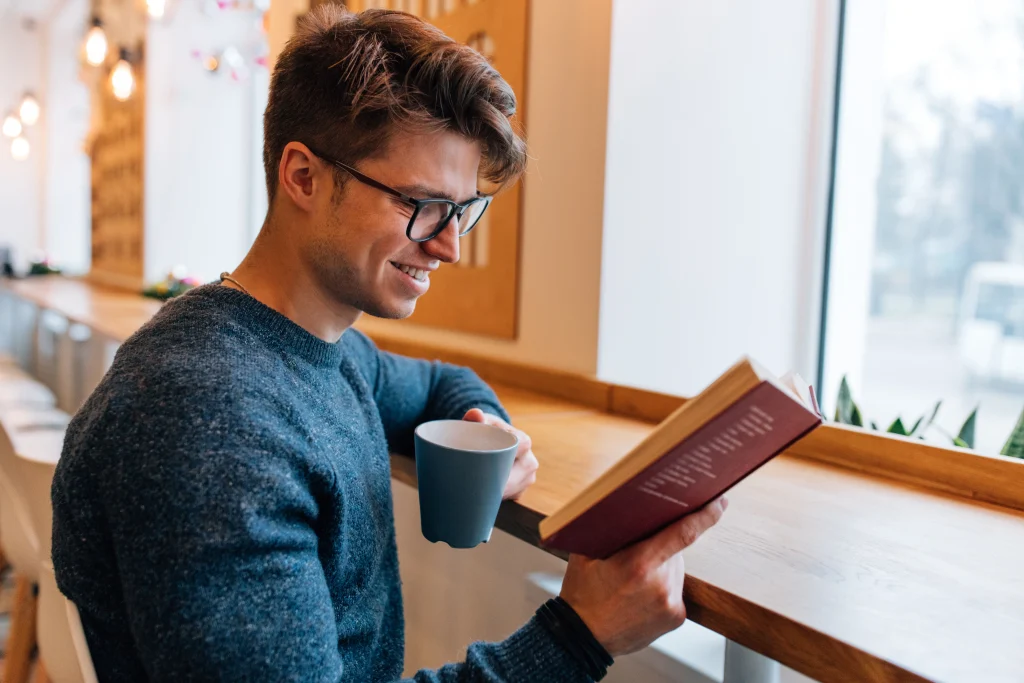 A smiling man with glasses sitting at a cafe counter, holding a coffee mug and reading a hardcover book near a window.