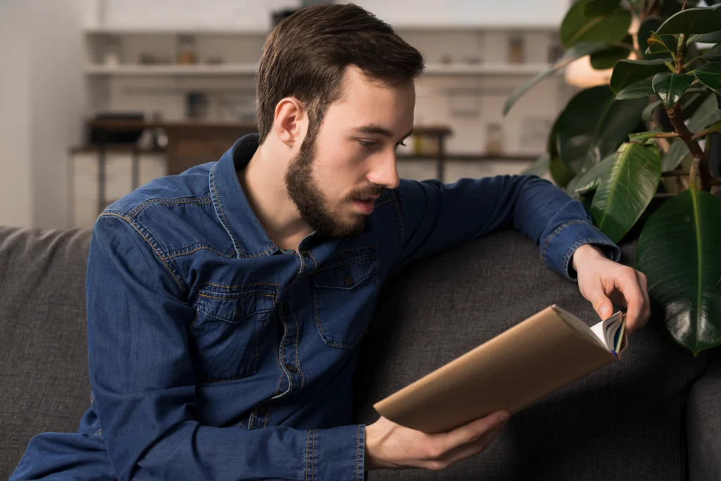 A man in a blue denim shirt sitting on a dark grey sofa, leaning forward to read a book while resting his arm on the back of the couch.