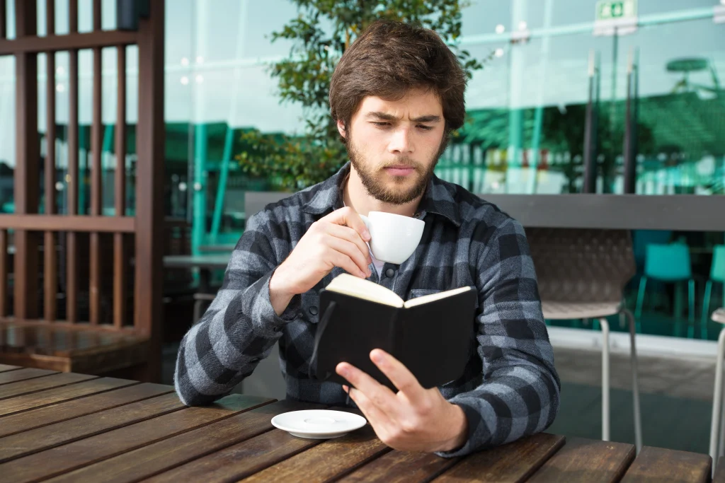A man with a beard sitting at an outdoor cafe table, holding a small white coffee cup while looking intently at an open black notebook.